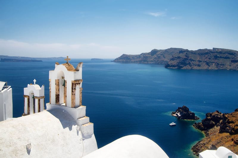 A white bell tower in Oia above the caldera, with the sea and the volcano islands behind