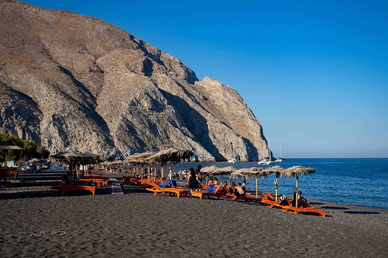 The black sand of Perissa beach with its rows of umbrellas and sunbeds, below the rock of Mesa Vouno